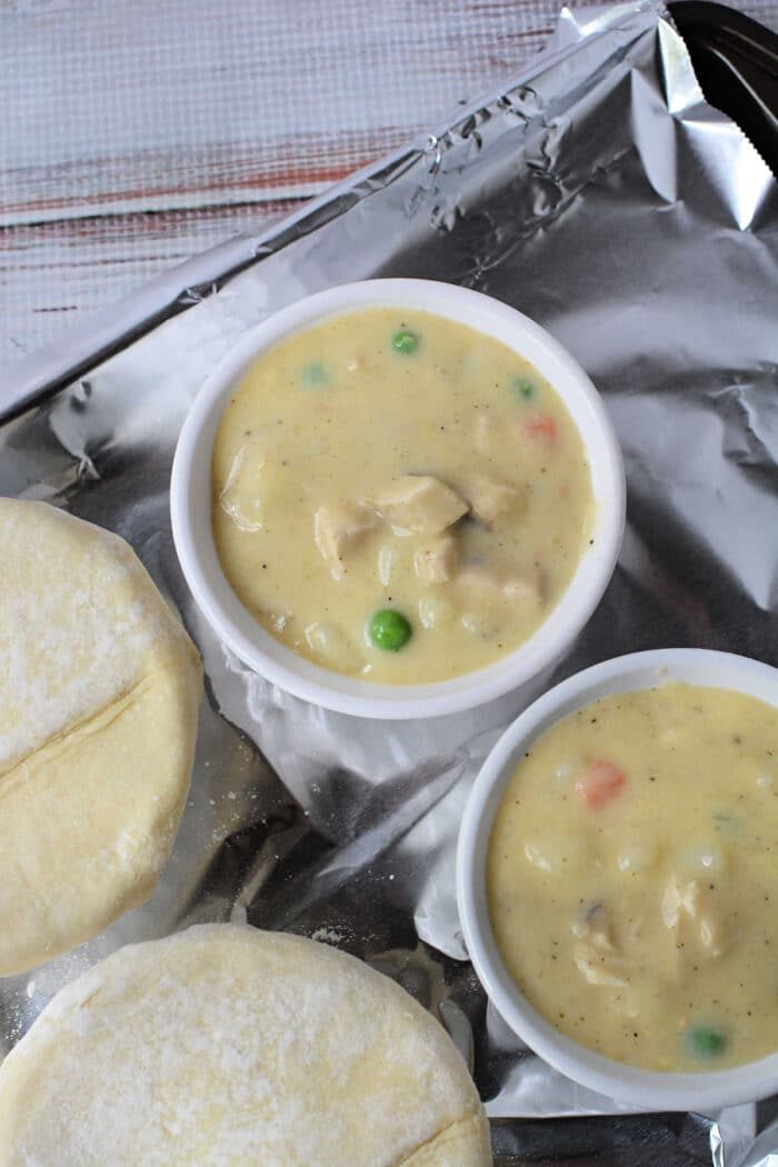 Two ramekins filled with creamy Chicken Pot Pie filling, next to uncooked pastry rounds on a foil-lined tray—a cozy recipe reminiscent of KFC's classic comfort food.