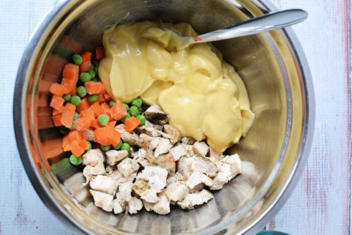 A mixing bowl with chopped chicken, frozen veggies, and creamy soup, like in a classic KFC Chicken Pot Pie, with a spoon inside.