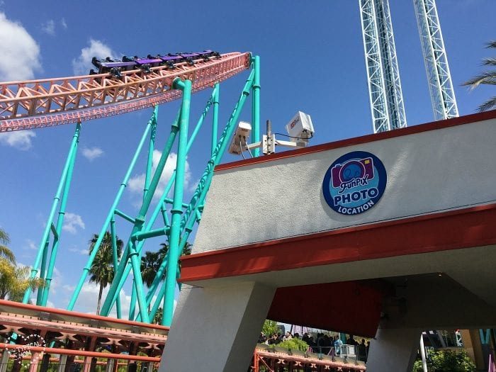 Roller coaster ascending on a turquoise track with riders under a blue sky at Knott's Berry Farm. A building below proudly displays a "Photo Location" sign, solid for capturing those FunPix memories.