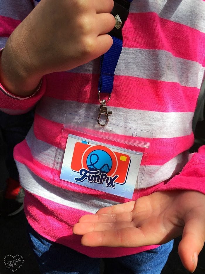 A child in a striped pink shirt holds an empty plastic badge holder attached to a lanyard featuring the Knott's Berry Farm FunPix logo in vibrant colors.