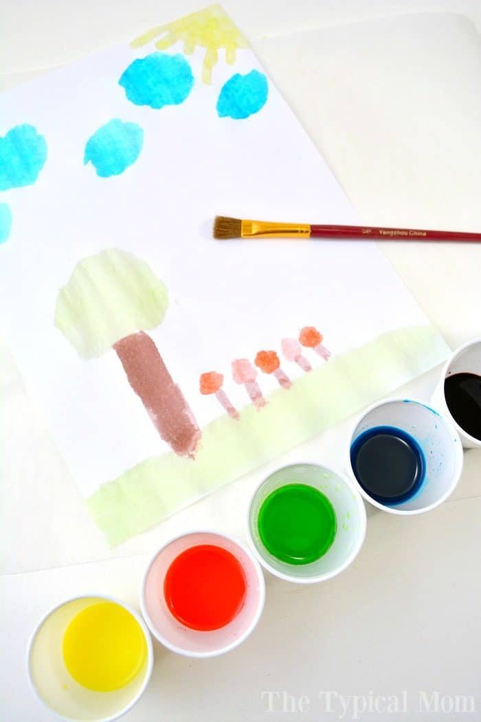 A child's watercolor painting of trees, sun, and clouds bursts with vibrant hues, reminiscent of a kool-aid paint palette. Nearby sit five small paint cups and a brush, ready for the next masterpiece.