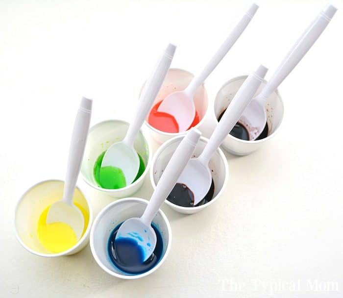 Six spoons rest in white cups filled with vibrant Kool-Aid-colored liquids: yellow, green, blue, red, teal, and purple, arranged on a white background.