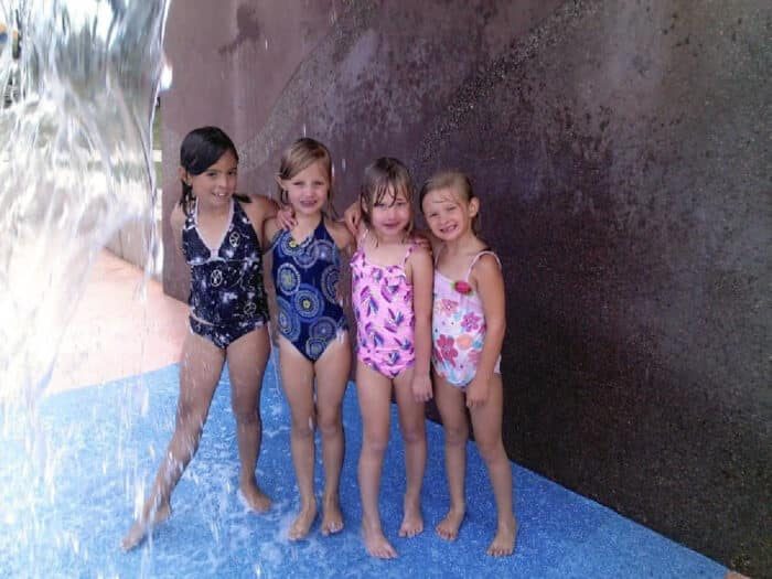 Four young children in swimsuits stand and smile under a water feature at what appears to be the Lake Skinner splash pad. A water stream is visible on the left, with the kids standing on a blue surface against a textured wall.