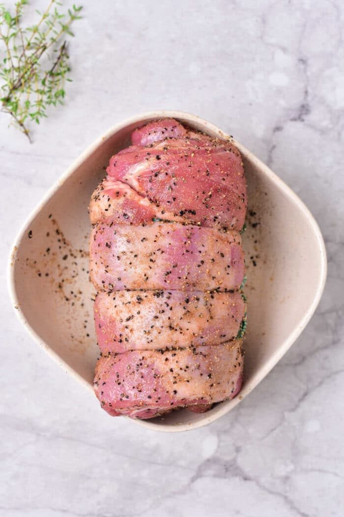 Raw seasoned rolled lamb breast in a white dish, primed for cooking, rests on a marble surface with a sprig of thyme nearby—a scene reminiscent of an exquisite lamb breast recipe.
