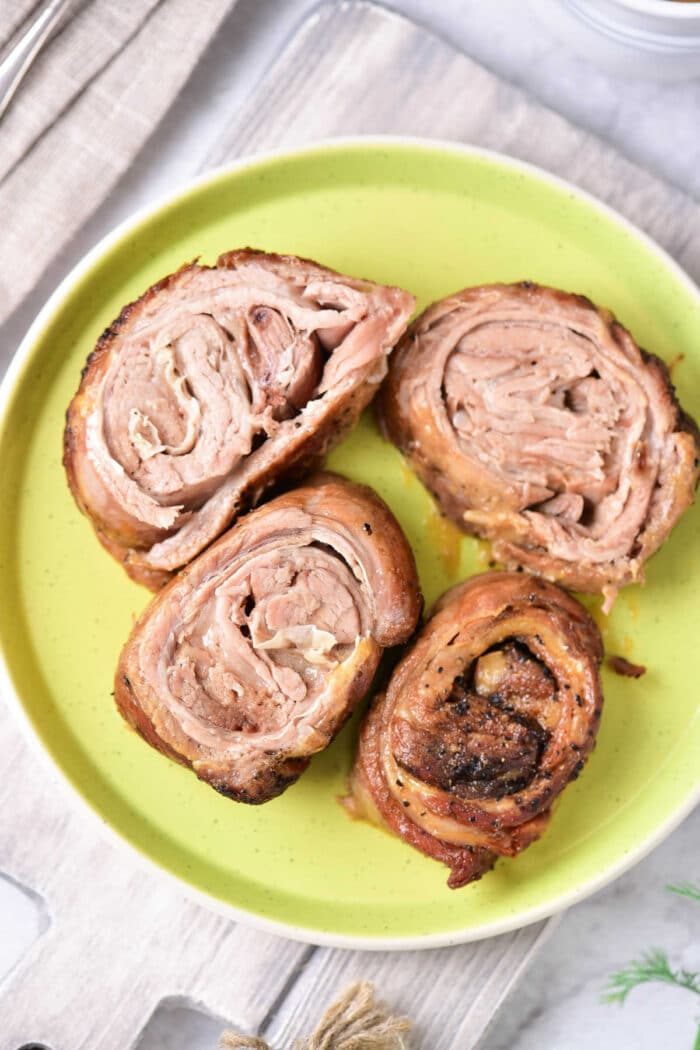 Four slices of cooked, rolled lamb rest enticingly on a green plate, reminiscent of a well-crafted lamb breast recipe.
