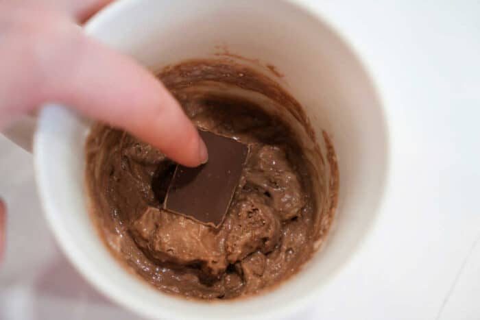 A hand presses a chocolate square into chocolate batter inside a white mug, preparing a quick and good Lava Cake.