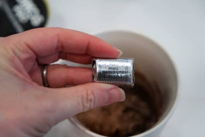 A hand holding a foil-wrapped Hershey's Nuggets chocolate above a cup of hot cocoa, solid for pairing with a slice of homemade chocolate banana bread.