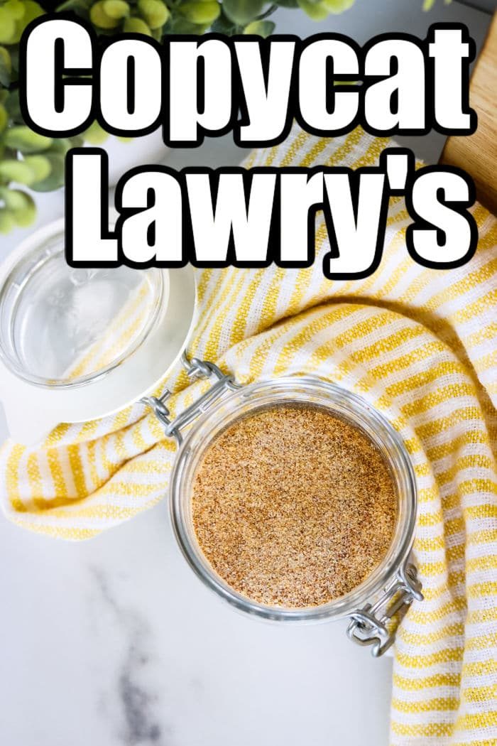 A glass jar filled with a seasoning blend sits on a white surface with a yellow striped cloth underneath. Above the jar, text reads "Copycat Lawry's Seasoned Salt Ingredients" in bold black and white letters. Greenery is partly visible in the background.