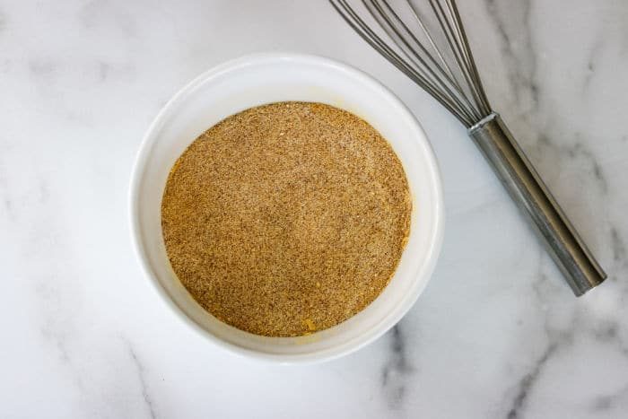 A white bowl filled with lawrys seasoned salt next to a metal whisk on a marble surface