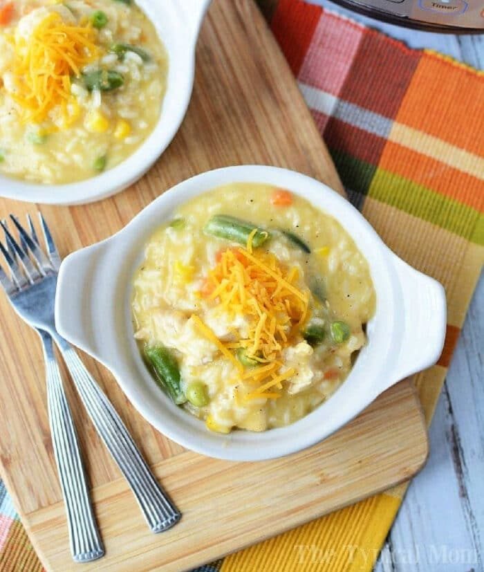 A bowl of creamy chicken and vegetables, crafted from leftover chicken breast, is topped with shredded cheese and sits invitingly on a wooden board alongside gleaming forks.