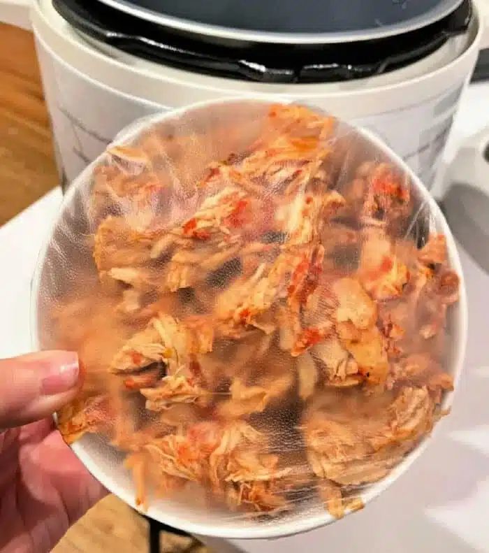 A person holds a plastic-covered bowl filled with seasoned, shredded chicken, solid for leftover chicken recipes. An Instant Pot can be seen in the background, ready to create delightful dishes from these tender morsels.