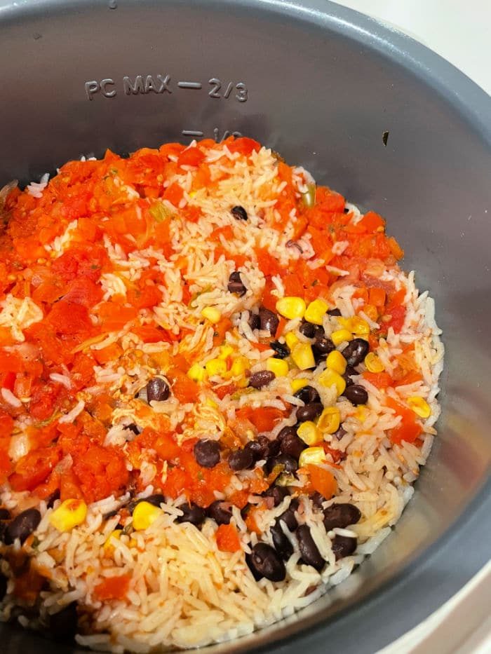 Rice, black beans, corn, and diced tomatoes are mixed together in an Instant Pot, with the option to add leftover chicken for extra flavor. The ingredients are partially cooked, showing distinct kernels of corn and tomato pieces. The cooker is marked with PC MAX – 2/3 on the side.