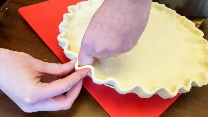 A person is crimping the edges of an unbaked leftover turkey pot pie crust in a metal dish. The dish sits on a red cutting board, and the hands are carefully shaping the scalloped edge of this cream-free creation.