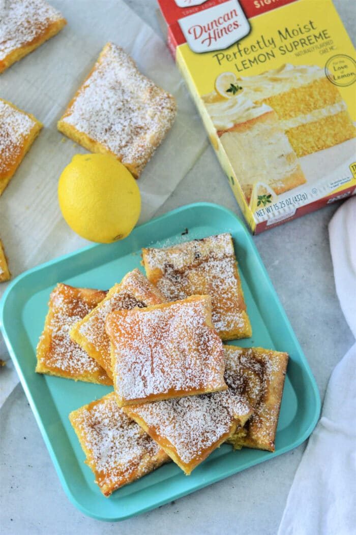 Lemon bars with cake mix dusted with powdered sugar rest elegantly on a blue tray, accompanied by a fresh lemon and the cake mix box for inspiration.