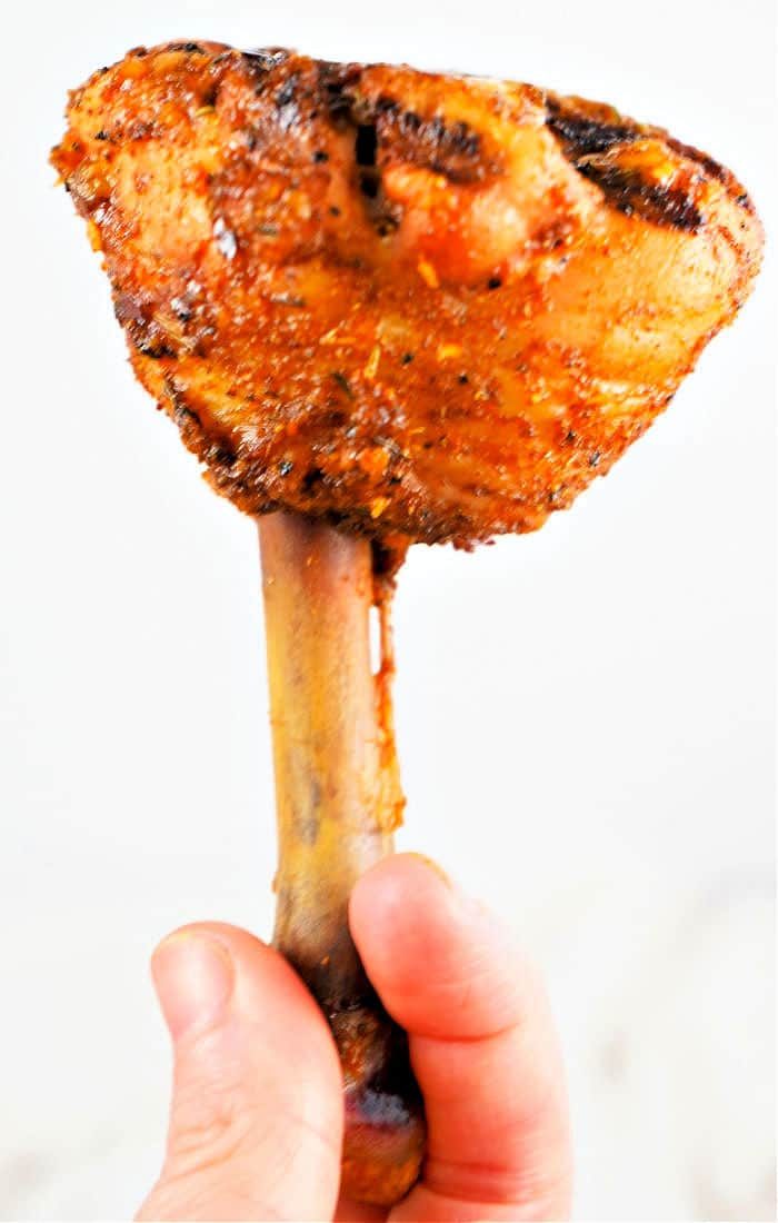A hand holds a perfectly seasoned and cooked giant lollipop chicken drumsticks, against a white background.