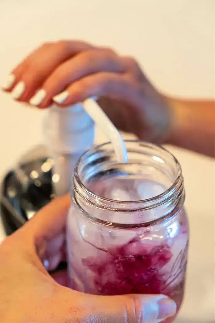 A person pumps liquid from a dispenser into a mason jar filled with a refreshing Lotus iced drink. The person's other hand carefully holds the jar while the pump's nozzle hovers above it, adding the solid touch. In the softly blurred background, hints of vibrant colors dance subtly out of focus.