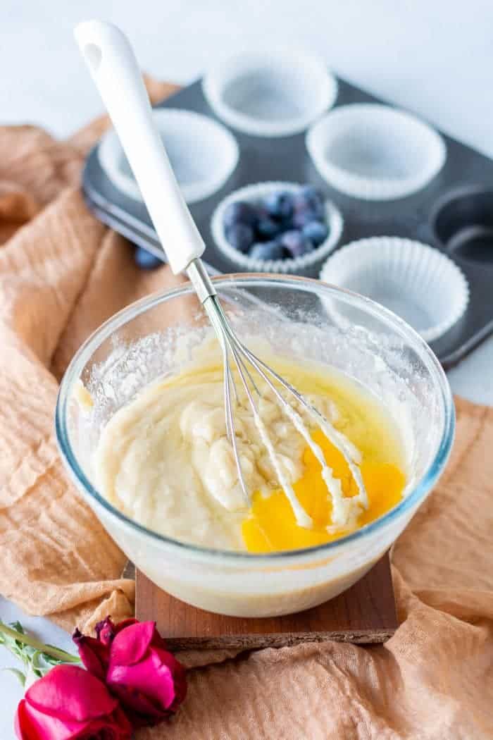 A glass bowl with muffin batter and whisk sits next to a muffin tray and rose on a beige cloth, ready for good frozen blueberry muffins.