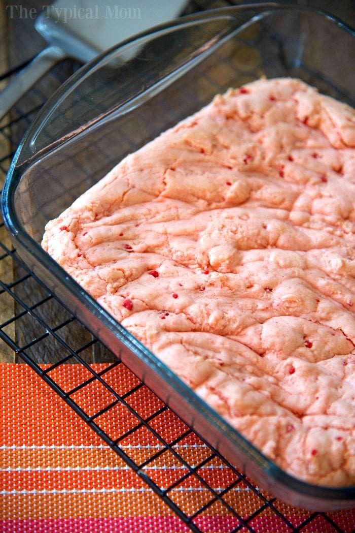 Strawberry brownie batter in a glass baking dish on a cooling rack atop an orange placemat hints at a simple twist: learn how to make brownies out of cake mix and surprise everyone with this vibrant treat.