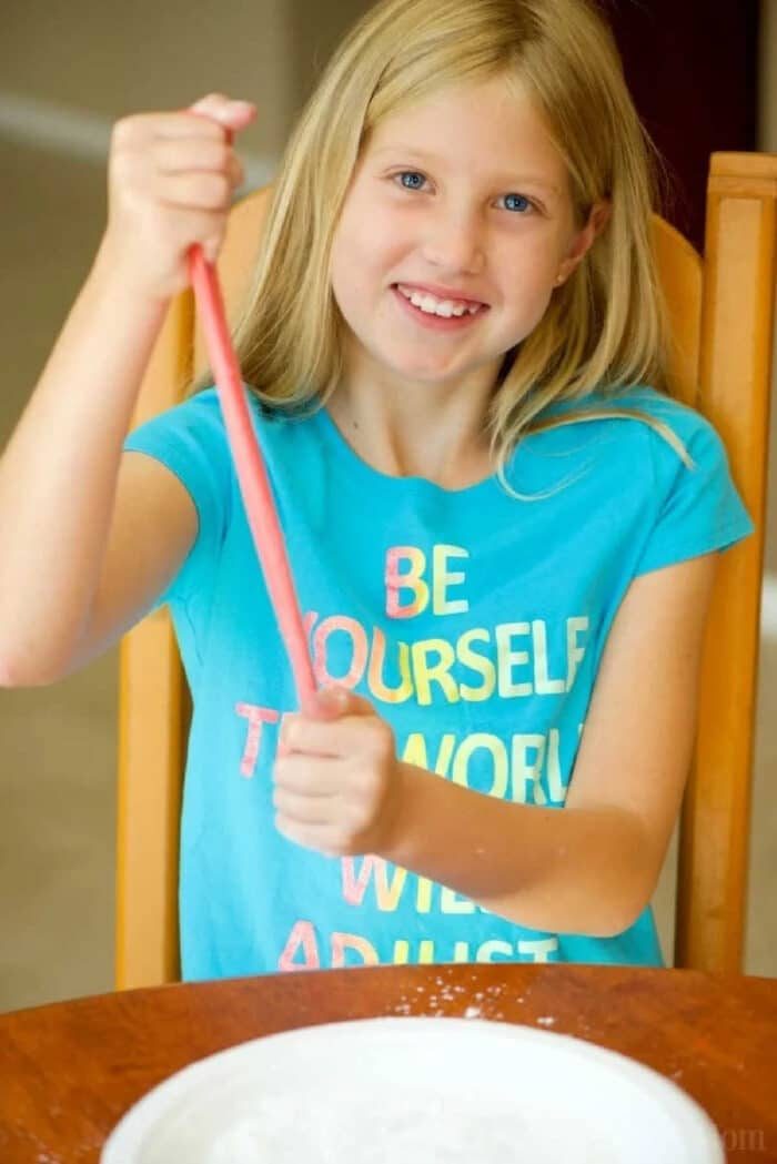 Smiling girl in a blue shirt stretches pink slime over a bowl while sitting at a table, showing how to make edible slime.