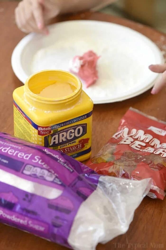 Open cornstarch container, powdered sugar bag, gummy bears, and a plate with pink dough on a wooden table—solid setup for learning how to make edible slime at home.