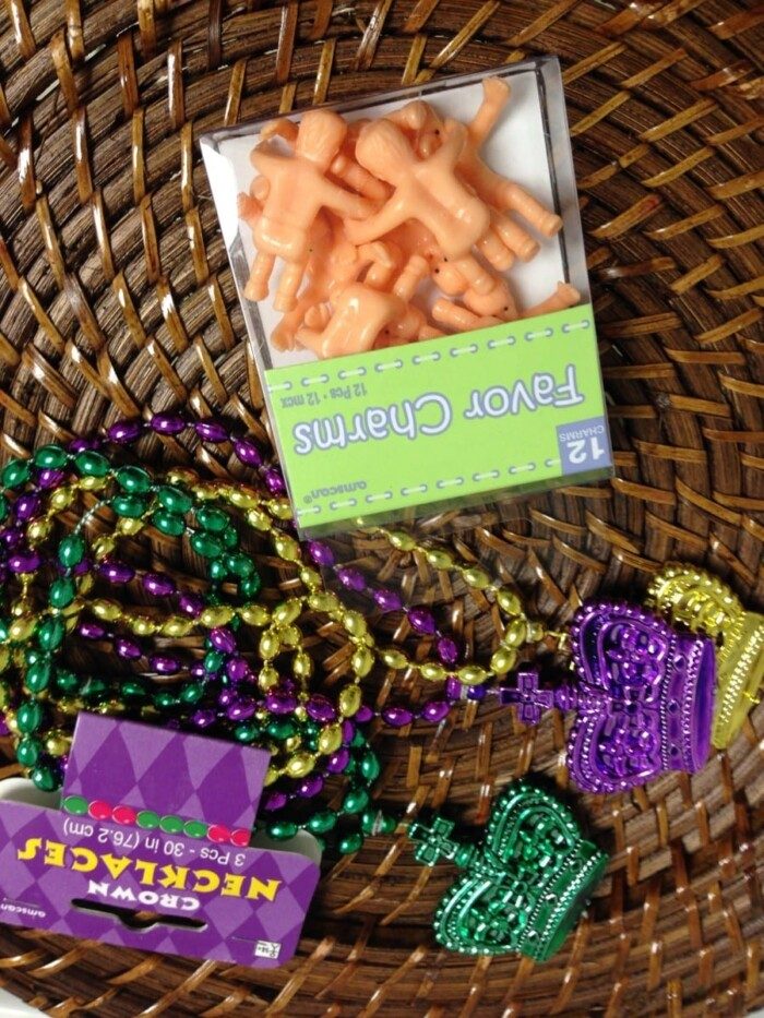A wicker basket holds colorful beaded necklaces in purple, green, and gold with crown and heart-shaped pendants. Nestled among them is a box of small plastic baby figurines labeled Favor Charms—solid for topping a Mardi Gras cake.