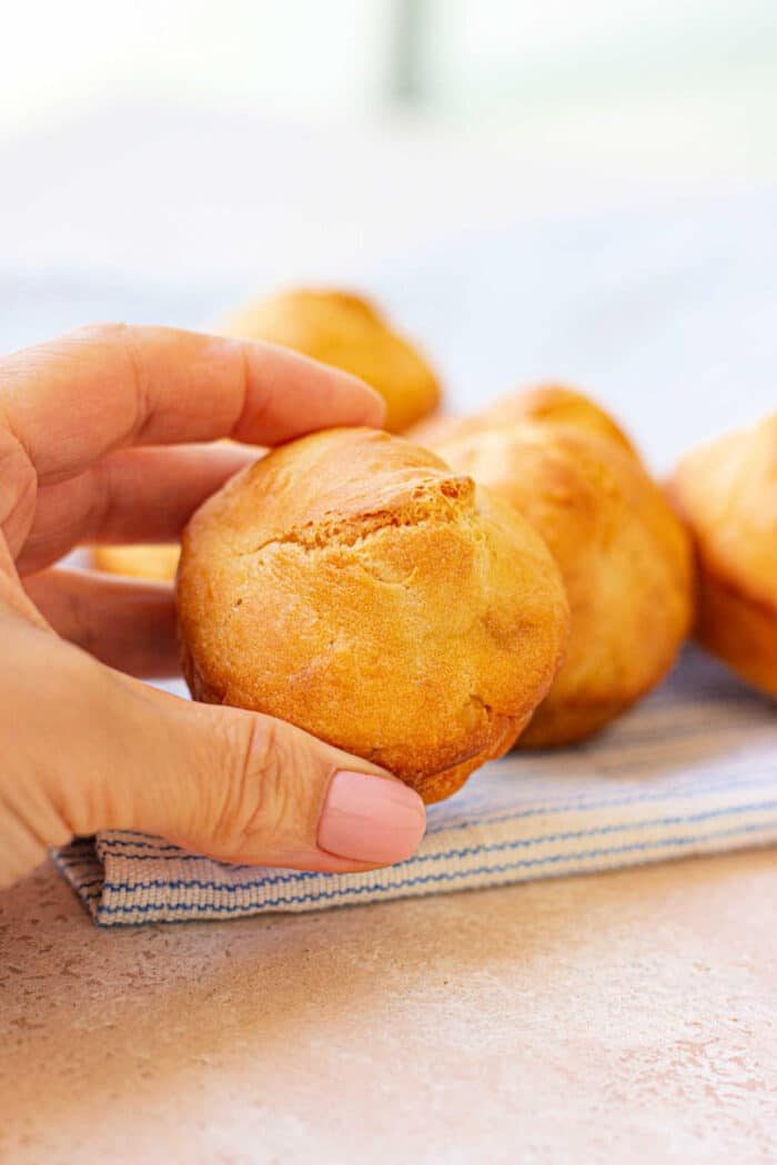 A hand holding a freshly baked mayo biscuit over a striped cloth resembles the warmth of homemade mayonnaise biscuits, with more muffins tempting in the background.