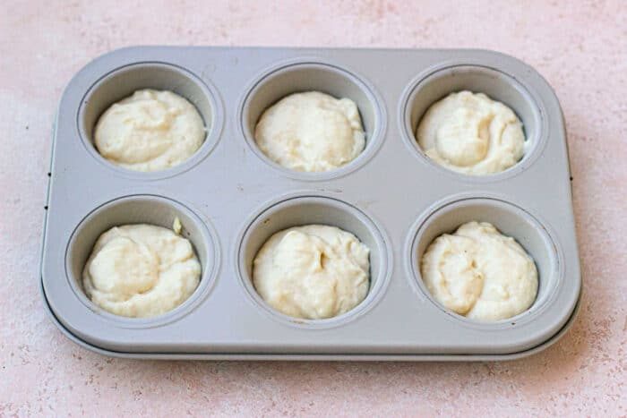 A muffin tin filled with six portions of creamy, unbaked batter sits gracefully on a light pink surface to make mayonnaise biscuits.