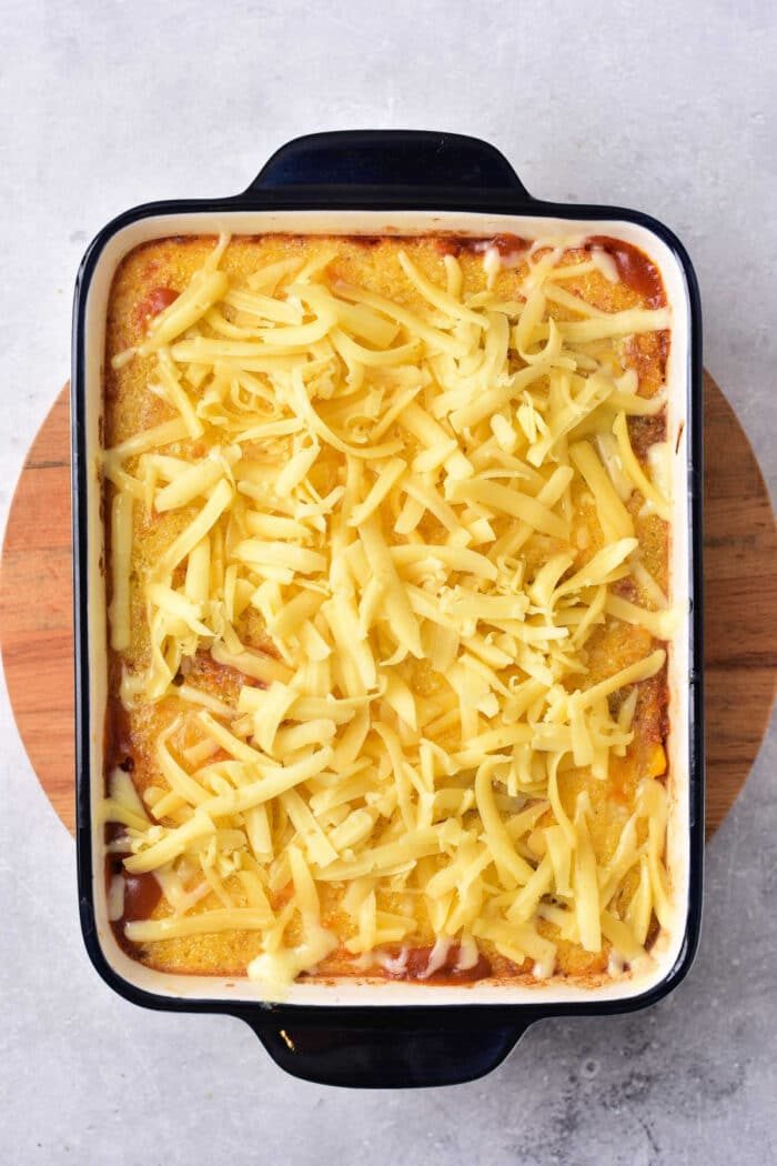 A wooden board cradles a casserole dish brimming with cornbread casserole, topped with golden melted cheese, reminiscent of a comforting Mexican cornbread casserole.