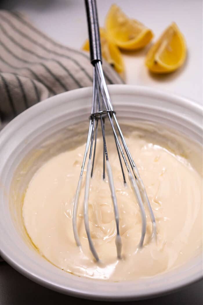 A whisk in a bowl of thick mayonnaise, with lemon wedges and a striped cloth in the background—its almost reminiscent of preparing Mexican mayonnaise.