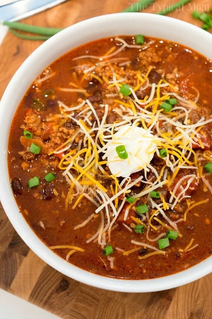 Bowl of ninja foodi chili topped with shredded cheese, sour cream, and chopped green onions on a wooden surface—a solid mild chili recipe for cozy nights.