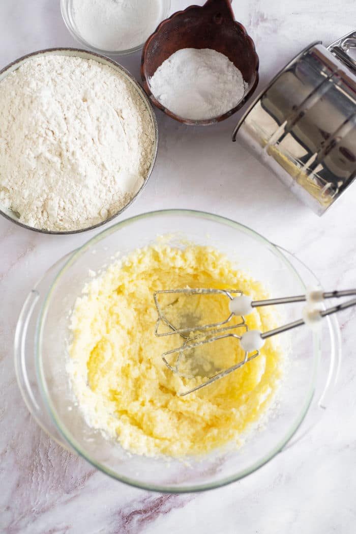 Bowl of creamed butter and sugar with beaters, surrounded by flour, sugar, baking powder for the solid start to a 3 Ingredient Pie Crust. A sifter rests nearby on the marble surface, ready to work its magic.