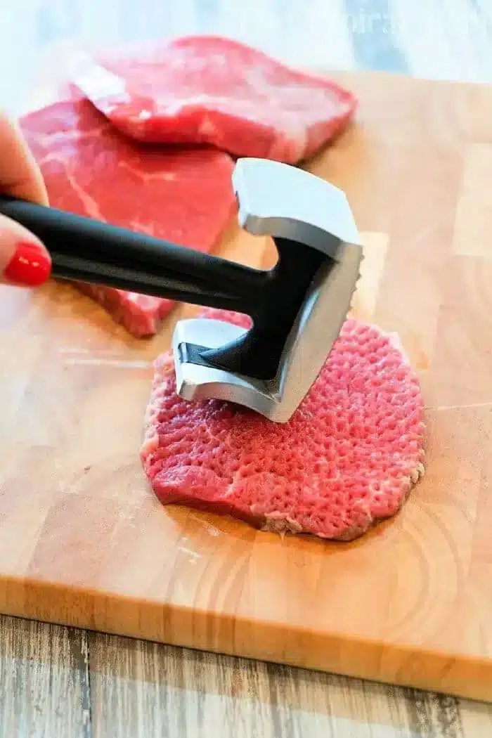 A person is expertly using a metal meat tenderizer on a piece of raw meat, preparing it for good minute steak recipes. The wooden cutting board holds the focus, while additional slices of meat patiently await their turn in the background.