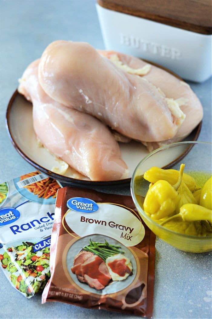 Raw chicken breasts on a plate with ranch mix, brown gravy mix, and a bowl of yellow peppers nearby—solid for making flavorful Mississippi chicken in the Instant Pot.