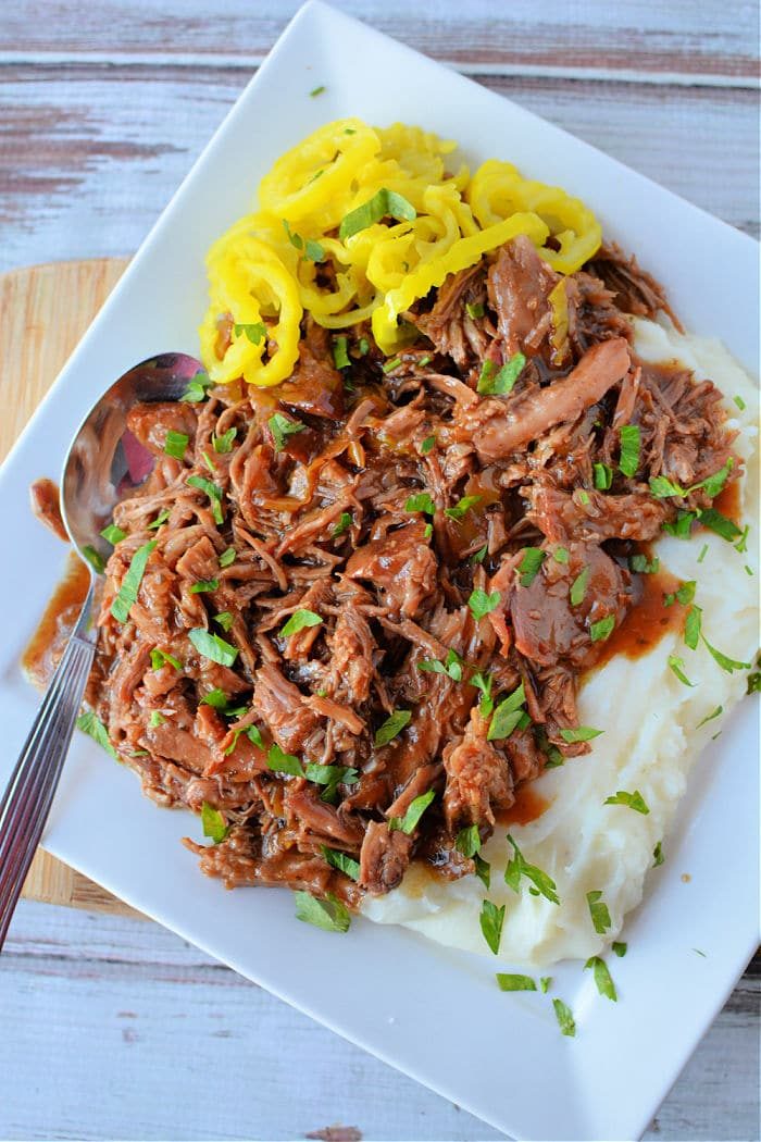 A white rectangular plate holds shredded pork with gravy, reminiscent of a Mississippi Pork Roast, garnished with chopped herbs next to a serving of creamy mashed potatoes. Slices of yellow pepper add a pop of color. A metal spoon rests on the plate's side.