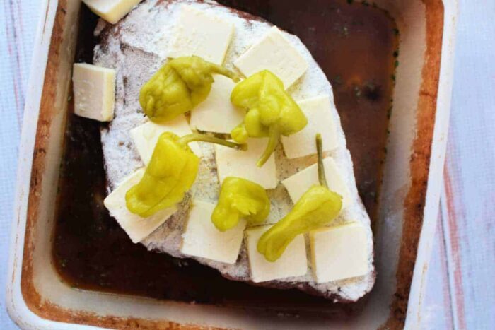 Mississippi Pot Roast Recipe featuring beef roast topped with butter, pepperoncini peppers, and seasoning in a baking dish ready to be cooked.