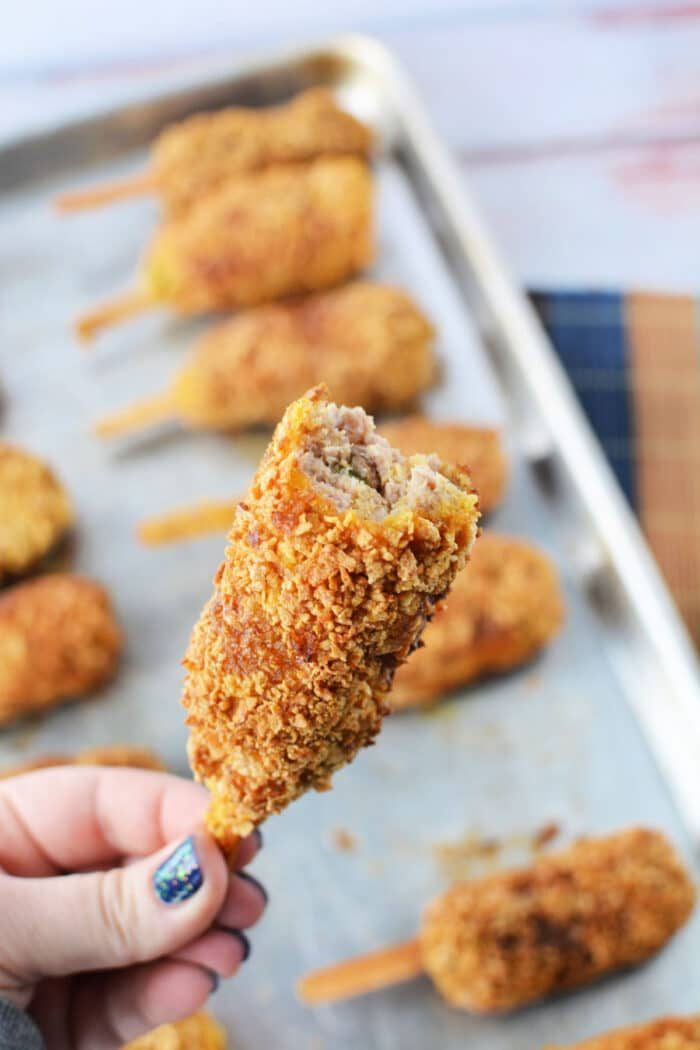 A hand holds a mock chicken leg with a bite taken out, revealing sausage inside. Nearby, mock chicken legs rest on a baking sheet, tantalizingly golden and ready to enjoy.