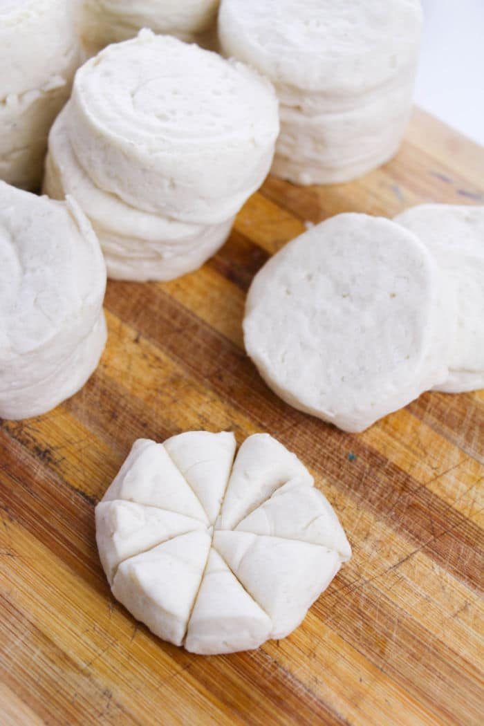 Stacks of uncooked biscuit dough, solid for making Monkey Bread, rest on a wooden board, with one sliced into eight pieces.