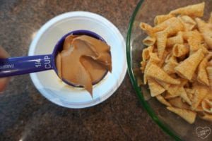 A measuring cup filled with peanut butter is held above a white container, ready to create some unicorn horn muddy buddies. Next to it, a green bowl holds a pile of rolled corn snacks, resting on the brown countertop in solid harmony.