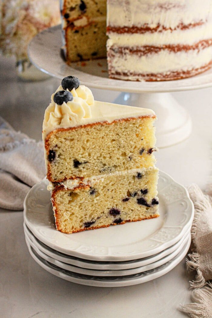 Slice of blueberry cake made with muffin mix, adorned with creamy frosting and a sprinkle of plump blueberries, resting on stacked plates. A whole cake sits invitingly in the background.