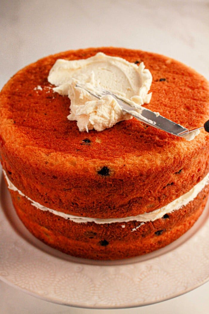 A delightful two-layered cake made with muffin mix, featuring a luscious cream filling. A knife gracefully spreads frosting on the top layer, all elegantly presented on a white plate.