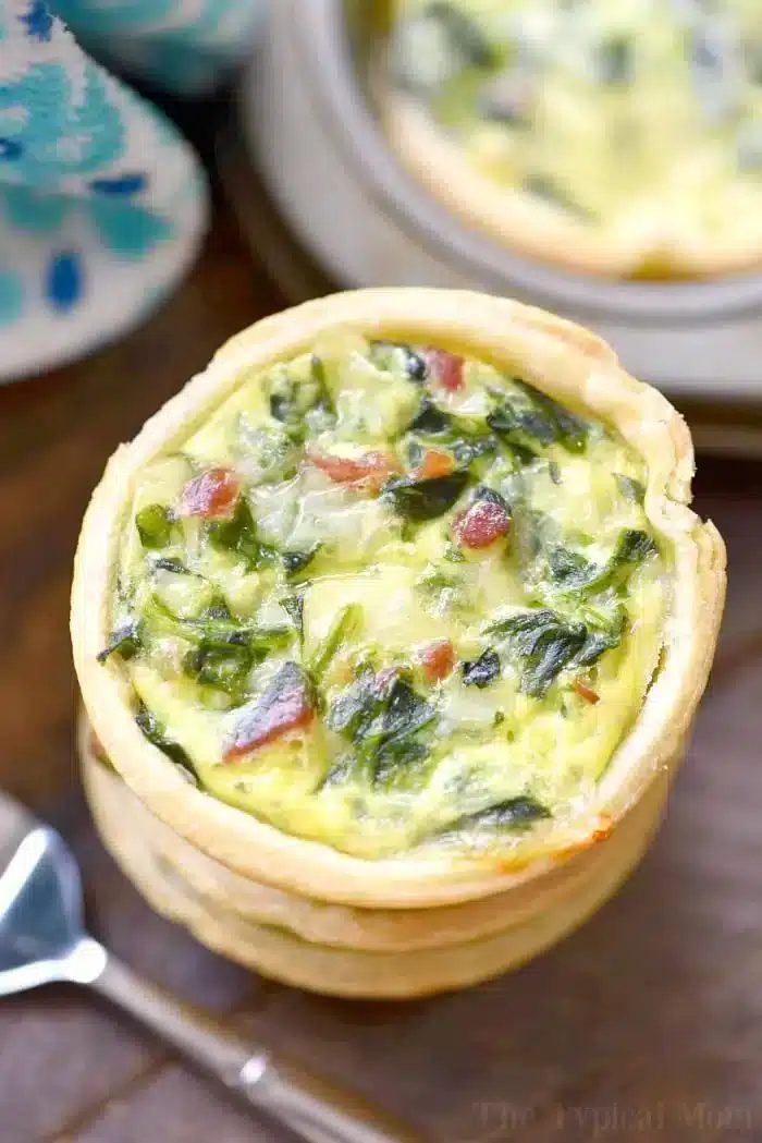 Close-up of a mini quiche with a golden crust, cooked to perfection using muffin tin recipes, filled with a savory mixture of eggs, cheese, spinach, and bacon pieces. The background includes a small plate and a spoon.