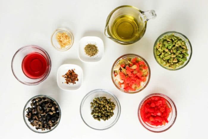 Various small bowls with chopped vegetables, olives, capers, spices, oil, and vinegar arranged on a white surface—solid ingredients for a classic Muffuletta recipe or homemade olive salad.