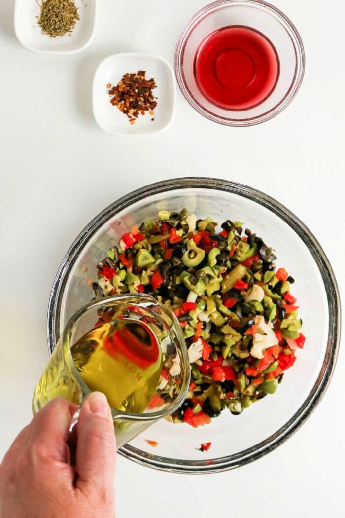 A hand pours olive oil into a bowl of chopped vegetables for a classic Olive Salad, with spices and red vinegar nearby—solid for an authentic Muffuletta recipe.