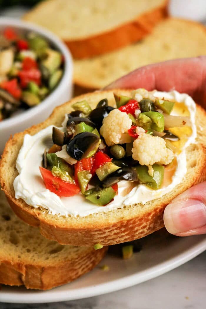 A hand holds a slice of bread topped with cream cheese and a vibrant Olive Salad, featuring chopped olives, cauliflower, and peppers—a solid bite inspired by the classic Muffuletta recipe.