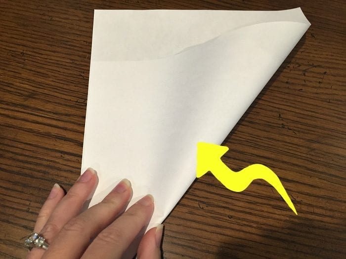 A hand is carefully folding a piece of white paper diagonally on a wooden surface, with a yellow arrow guiding the fold direction, setting the stage for crafting a multiplication fortune teller game.