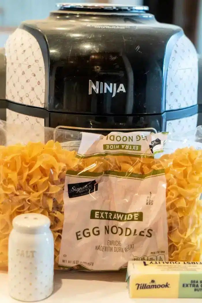 A sleek black Ninja Foodi air fryer looms in the background. Front and center, you'll find packages of egg noodles, a block of creamy Tillamook butter, and a chic white salt shaker adorned with gold dots—solid ingredients for crafting delightful butter noodles.