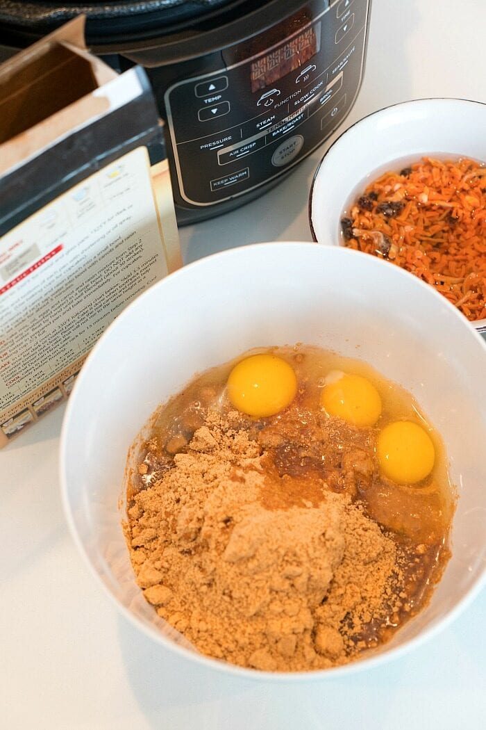 A bowl with eggs and brown mix sits alongside a Ninja Foodi cake, with an Instant Pot and various ingredients scattered in the background on a kitchen counter.
