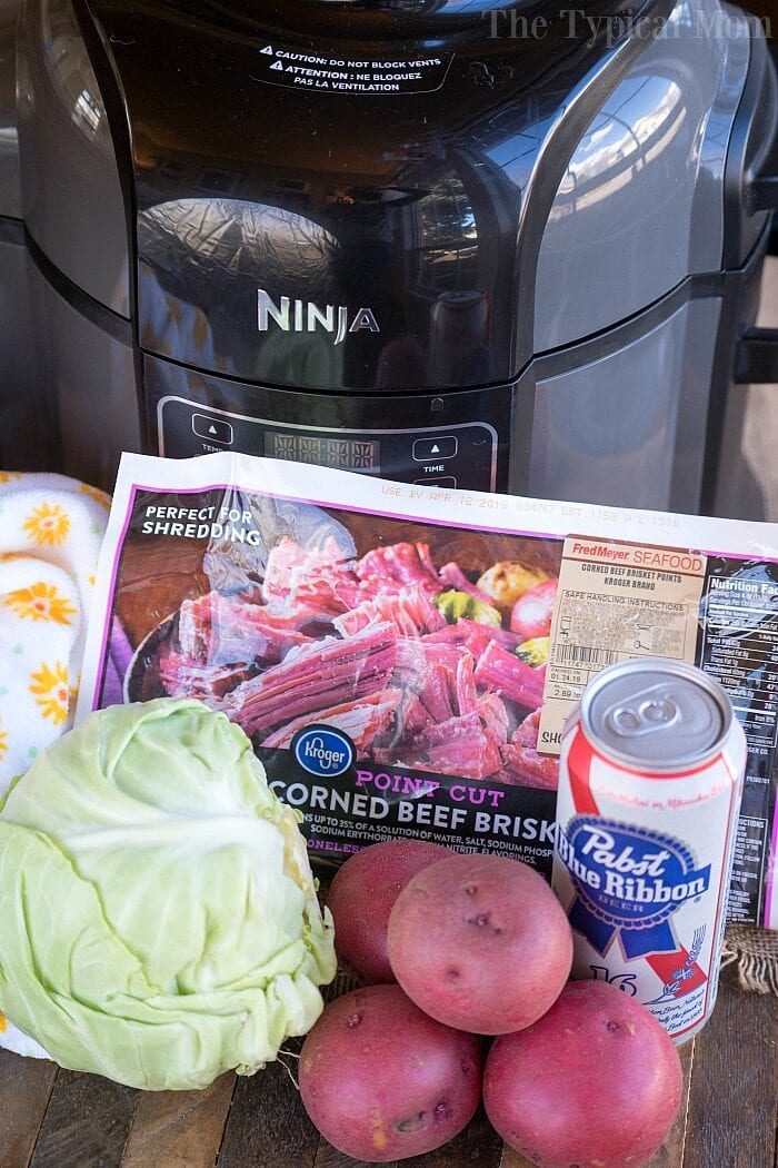 On a wooden table sits a tempting array: cabbage, red potatoes, canned beer, and a corned beef brisket package. solid for creating the ultimate Ninja Foodi corned beef and cabbage feast.