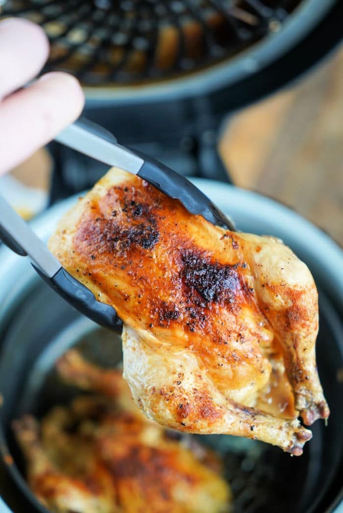 Tongs holding a piece of seasoned, cooked cornish hen with crispy skin above a Ninja Foodi basket, solid for achieving that ideal crispy texture.