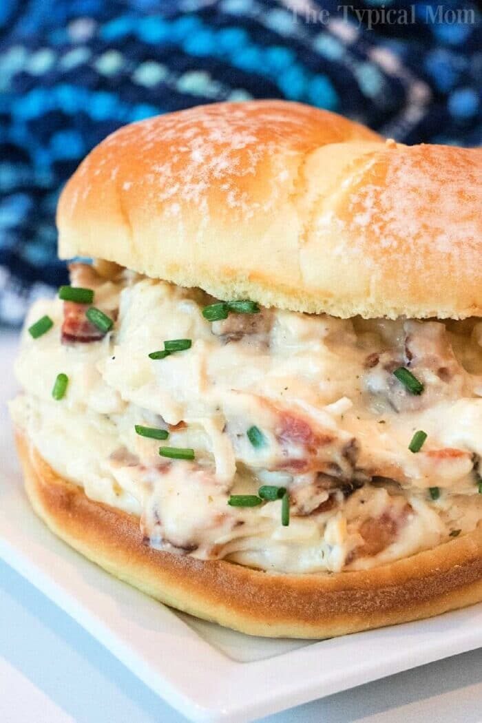 A creamy crockpot crack chicken bacon sandwich on a toasted bun, garnished with chopped chives, served on a white plate with a blue patterned cloth in the background.